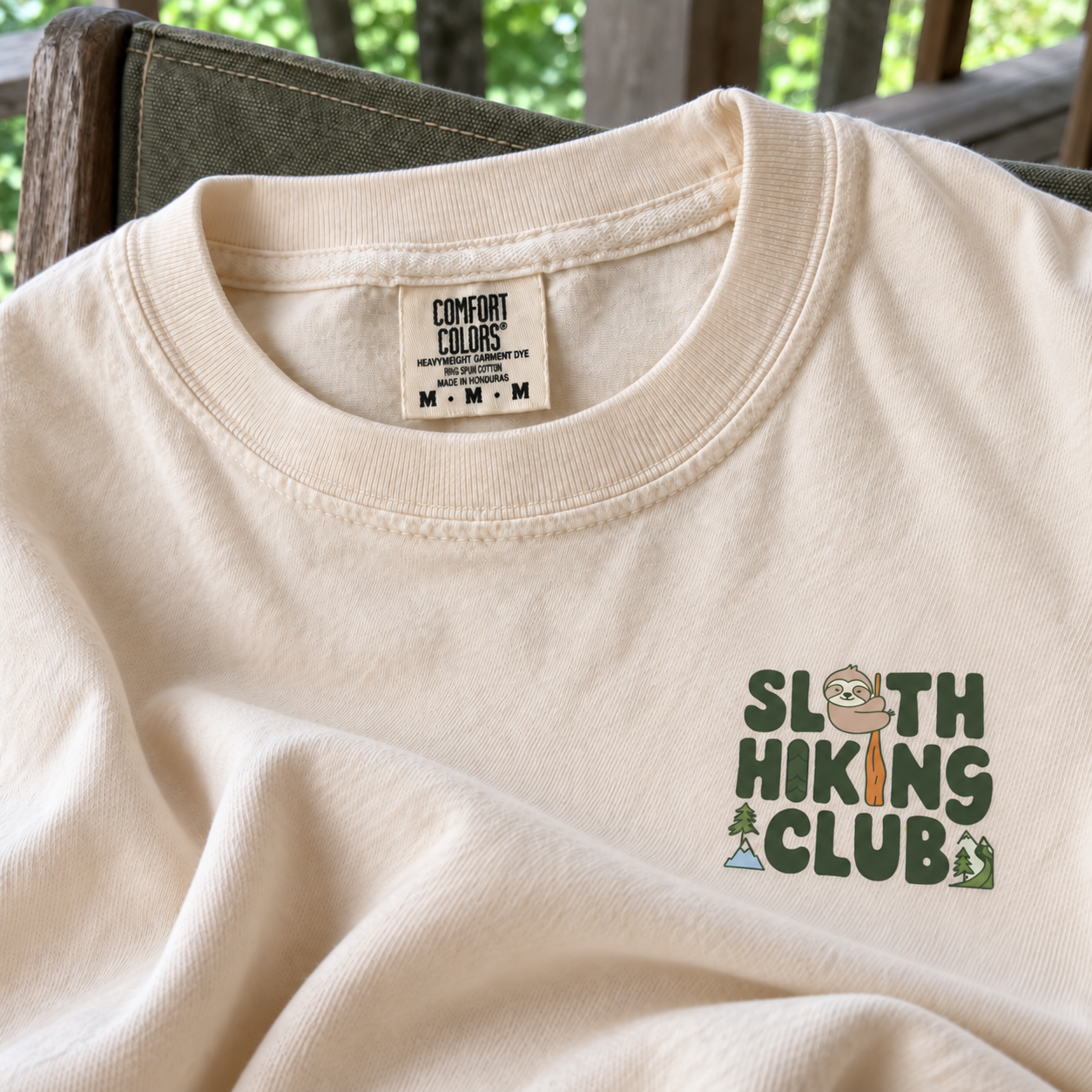Close-up of an ivory Sloth Hiking Club Comfort Colors tee on an outdoor chair, showing its cotton texture, stitched collar, neck label and small chest print.